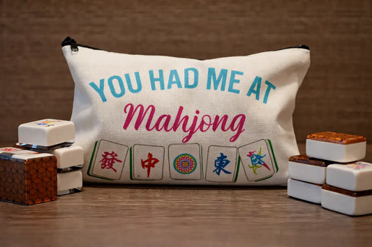 White canvas tile bag with “You Had Me at Mahjong” text and printed mahjong tiles, displayed with stacked game tiles on a wooden surface.