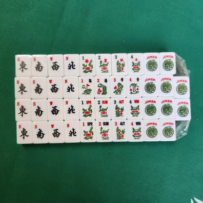 A set of Mahjong tiles arranged in rows on a green felt surface, with some tiles still wrapped in clear plastic.