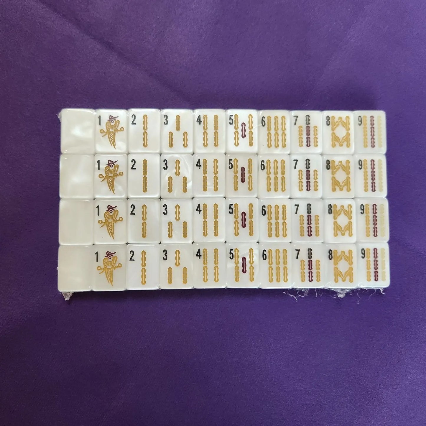 A set of white mahjong tiles arranged in a grid on a purple fabric background. The tiles feature gold-colored Chinese numerals and traditional symbols.