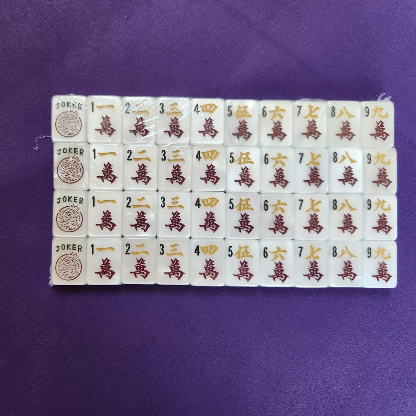 A set of Mahjong tiles arranged in a grid on a purple background. The tiles are white with gold and red Chinese characters and symbols, including Joker tiles on the left.
