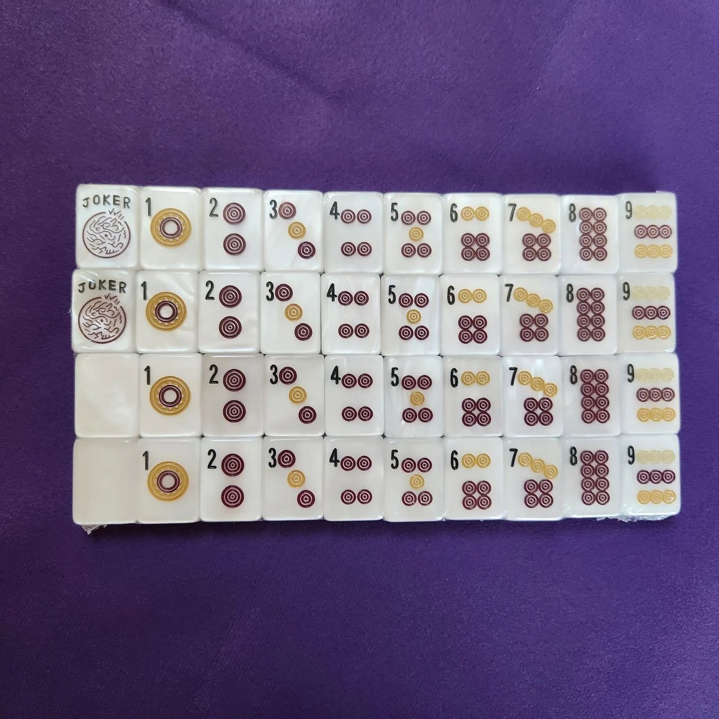 A set of Mahjong tiles arranged in rows on a purple fabric background. The tiles are white with printed symbols in dark red, gold, and black, including Joker tiles with a circular emblem.