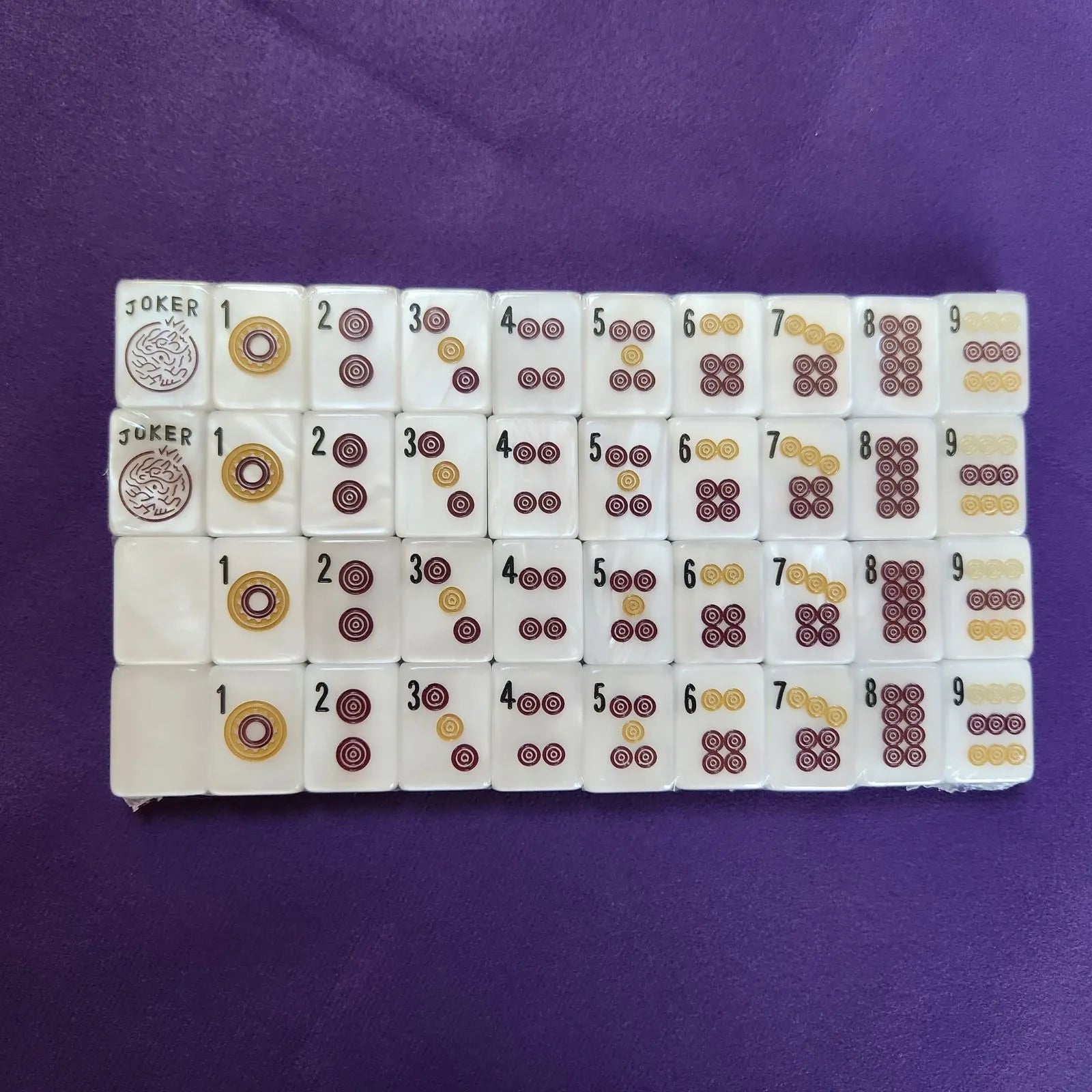 A set of Mahjong tiles arranged in rows on a purple fabric background. The tiles are white with printed symbols in dark red, gold, and black, including Joker tiles with a circular emblem.