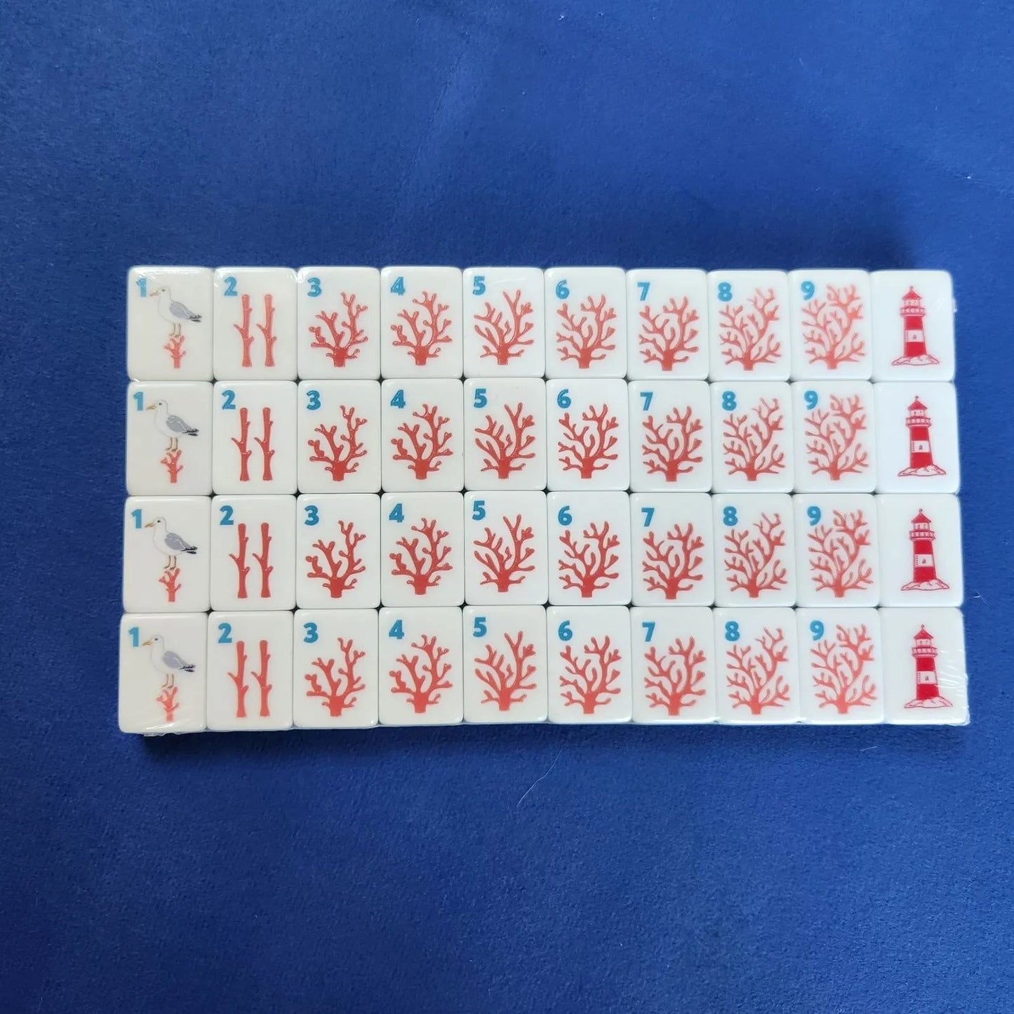 A set of Mahjong tiles arranged in a grid on a textured blue background. The white tiles feature red coral illustrations, blue numbers, and traditional Chinese symbols.