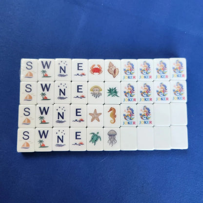 A set of white mahjong tiles arranged in a grid on a blue background. The tiles feature various sea-themed illustrations such as a crab, seashell, octopus, jellyfish, starfish, seahorse, turtle, sailboat, waves, and the words SW, N, E, W, JOKER.