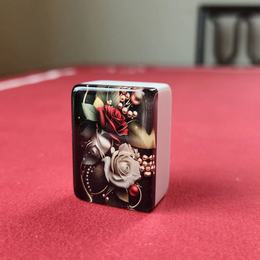 A square decorative object with rounded corners, featuring a glossy floral design with red and white roses, green leaves, and metallic gold accents on a dark background, standing on a red surface.