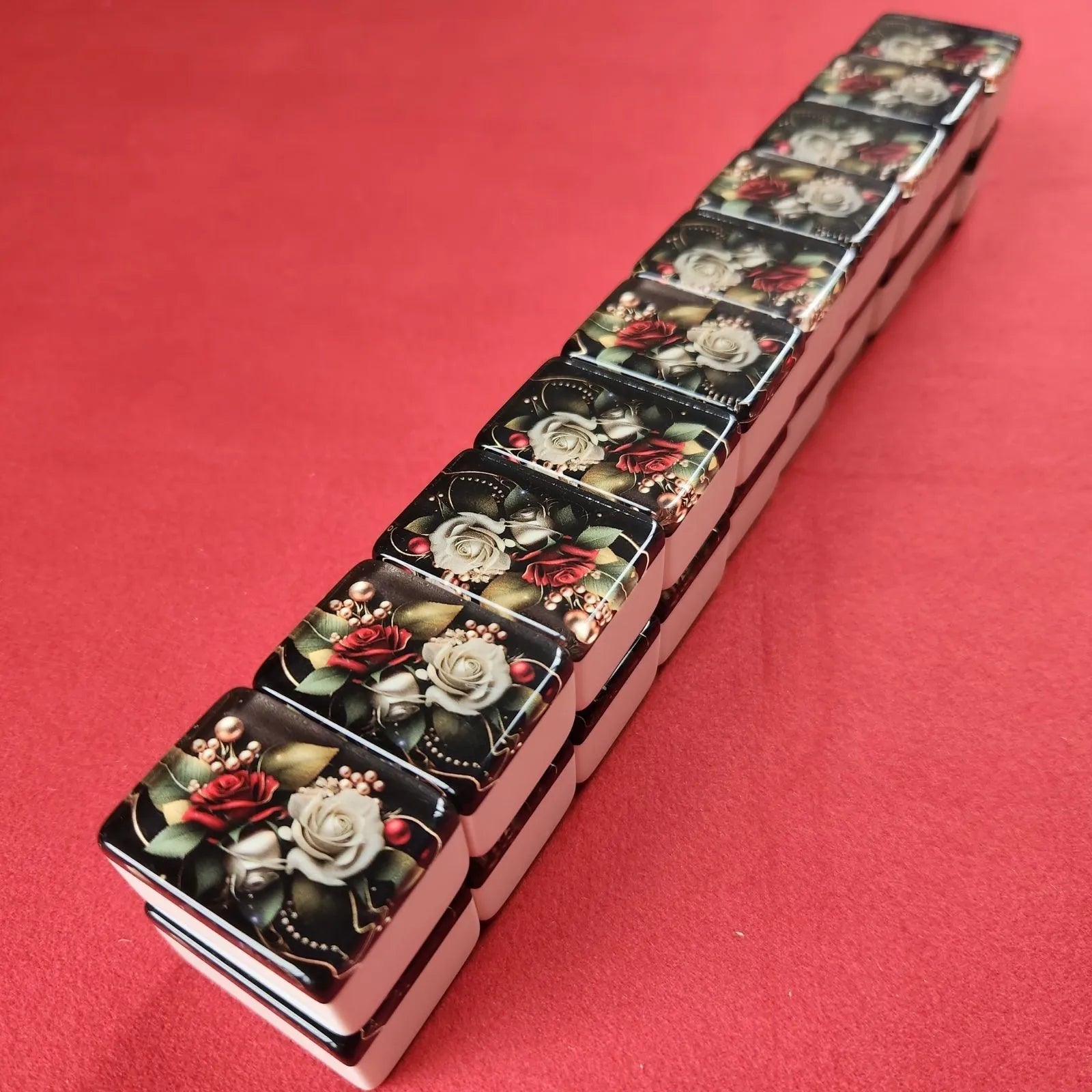 A set of mahjong tiles arranged diagonally on a solid red background. The tiles feature a glossy black background decorated with detailed illustrations of red and white roses, green leaves, and gold accents.
