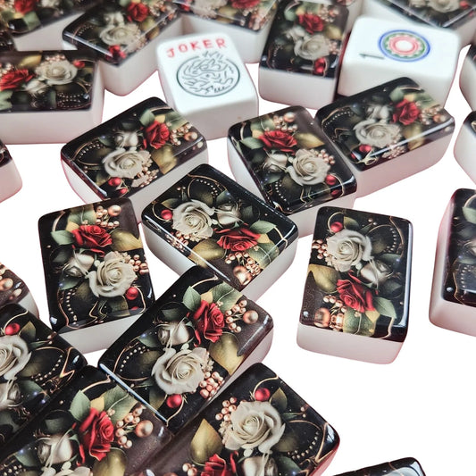 A close-up of a pile of mahjong tiles on a white background. Most tiles feature a black background with a detailed floral print of red roses, white roses, and green leaves.