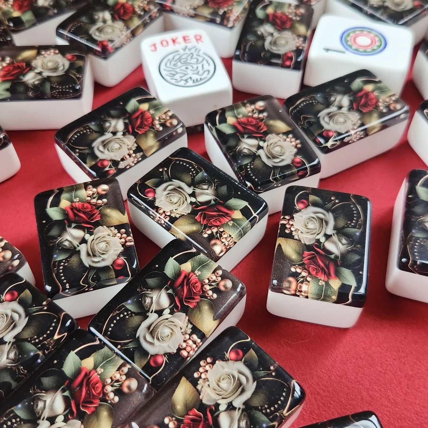 A pile of mahjong tiles with a glossy black back and white front faces, decorated with detailed illustrations of red roses, white lilies, green leaves, and gold accents. One tile is clearly visible with the word 'JOKER' printed on it.