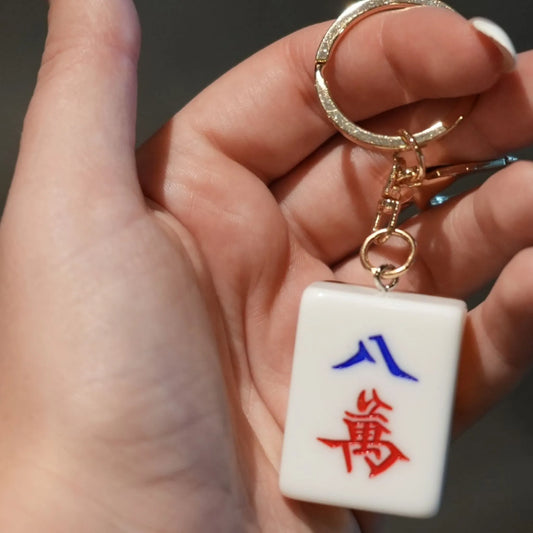 Hand holding a keychain with a Mahjong tile design featuring red and blue characters.