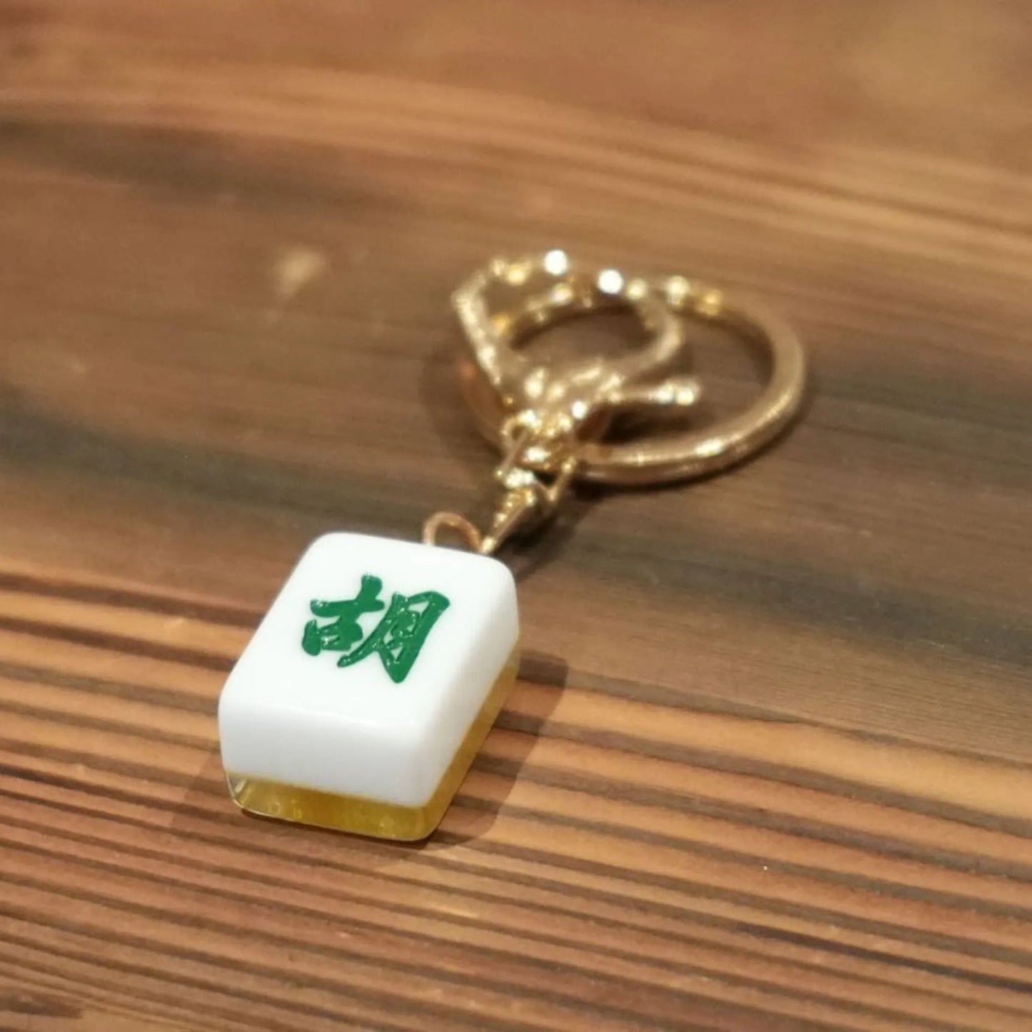 Mahjong keychain with green character on white tile, gold ring, on wooden surface